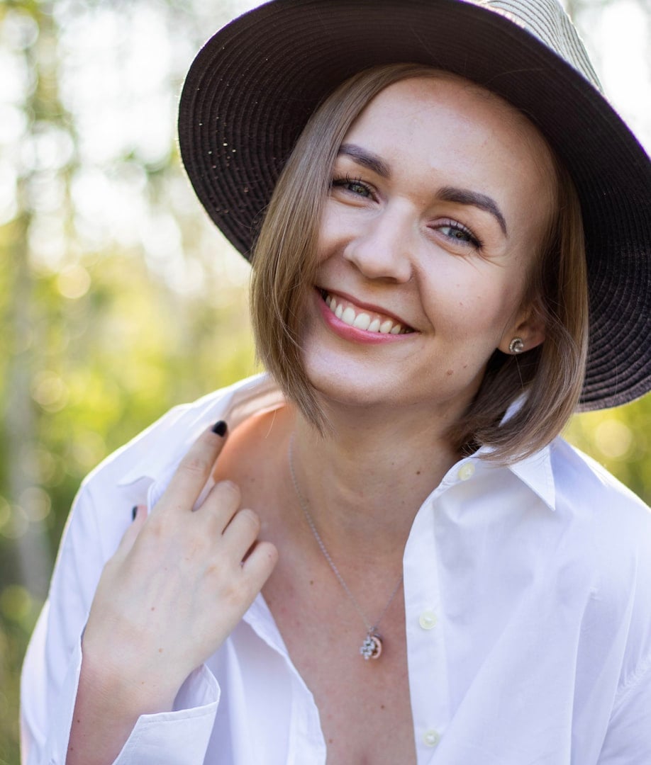A woman with short brown hair smiles, wearing a light blue t-shirt, against a soft-focus neutral background.